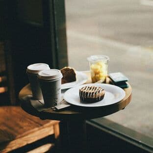 Two paper coffee cups, a muffin, and a cinnamon bun on a small round wooden cafe table by the window.