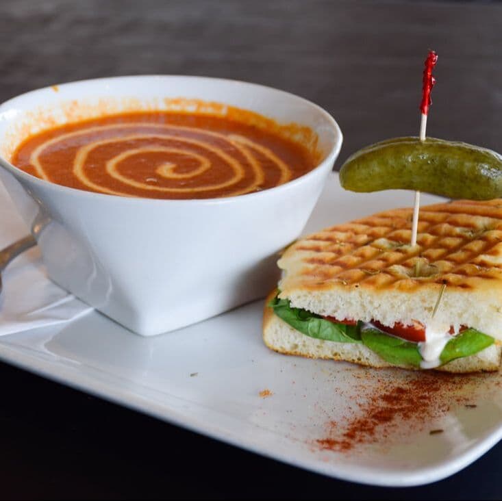 A bowl of Java Garden roasted red pepper and tomato soup with a tomato-mozzarella panini and a pickle.