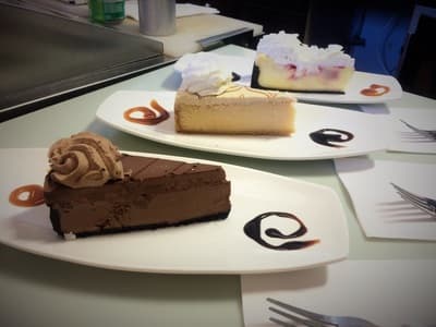 Three slices of cheesecake on white plates: chocolate, vanilla, and a strawberry-topped slice.