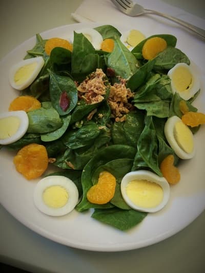 Spinach salad with hard-boiled egg, mandarin orange, and bacon crumble on a white plate.